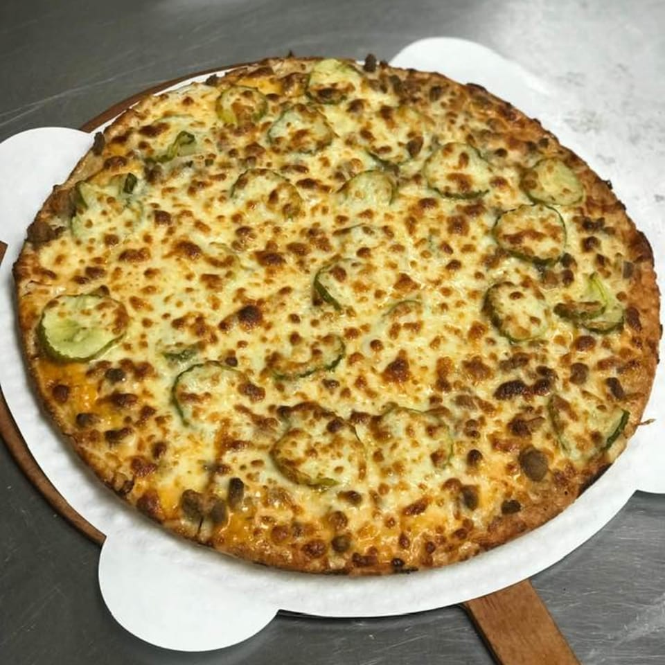 A Runner Pizza Stoughton pizza with melted cheese and pickle slices on a wooden peel.