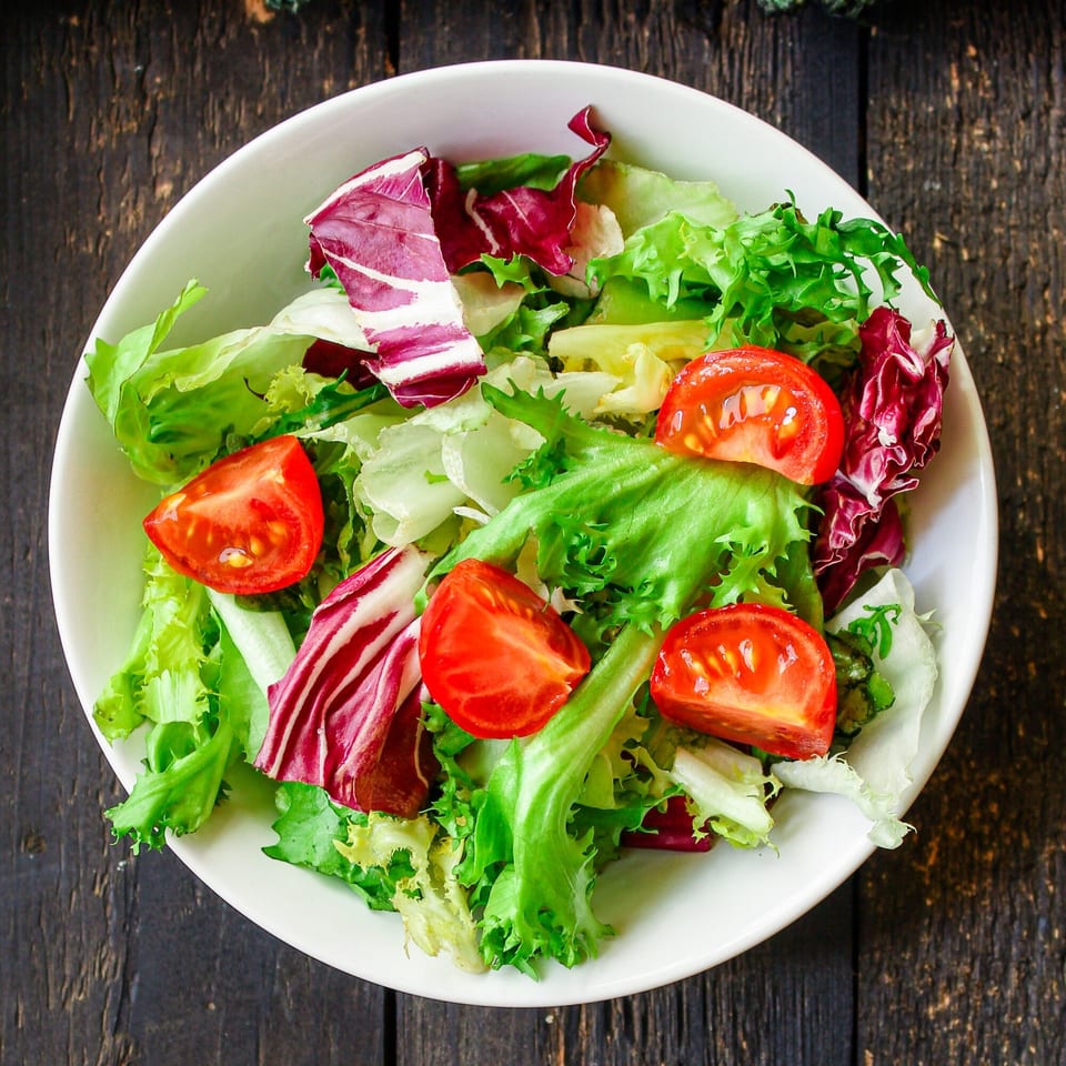Fresh garden salad with mixed greens and quartered tomatoes in a white bowl on a dark wooden table.