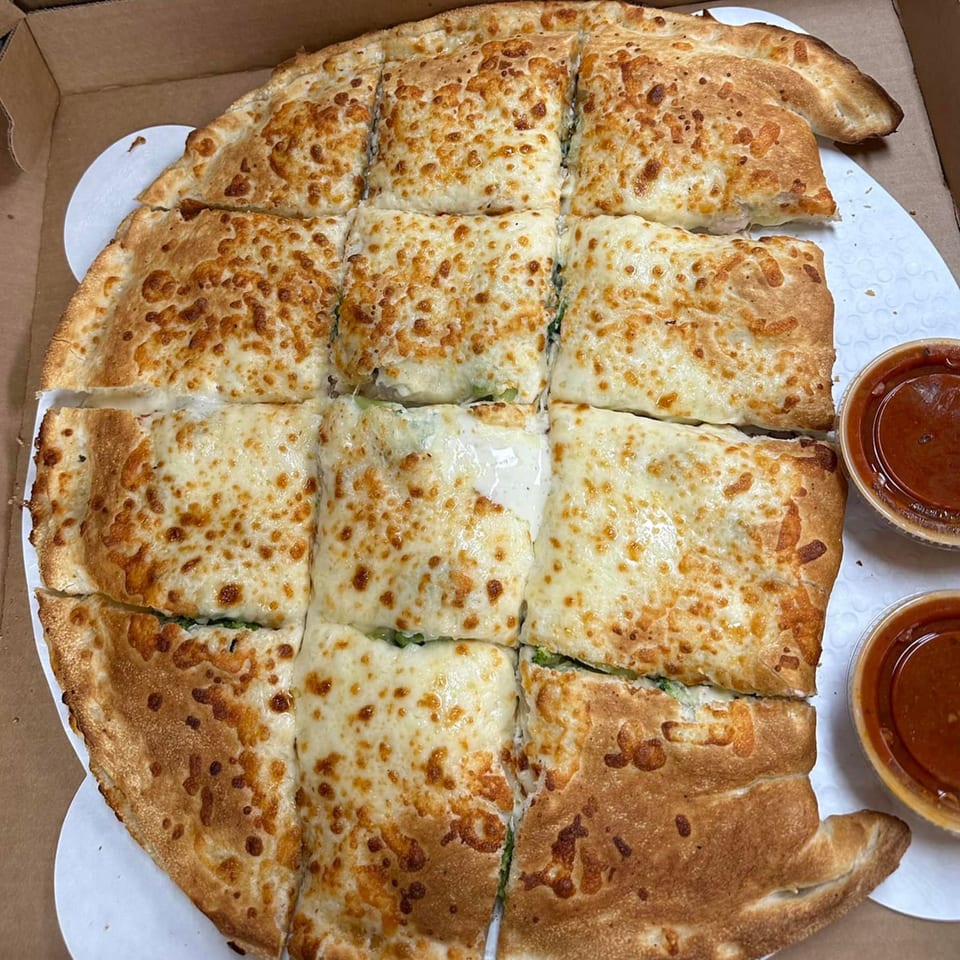 A large, cheesy calzone, cut into squares, with two small cups of marinara sauce in a pizza box.